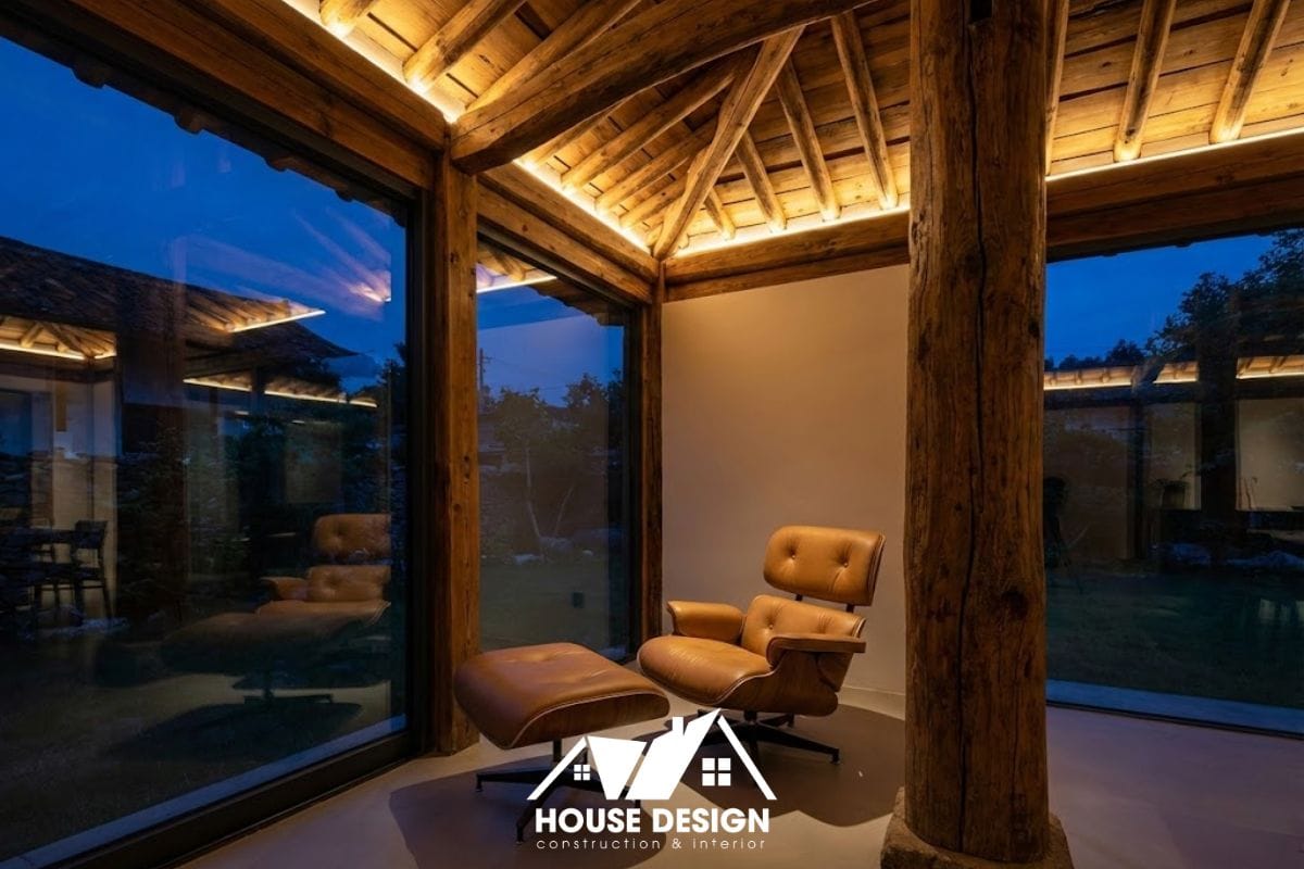 Góc đọc sách yên tĩnh trong nhà gỗ với ghế thư giãn và hệ thống đèn hắt trần hiện đại tôn vinh hệ kèo gỗ.